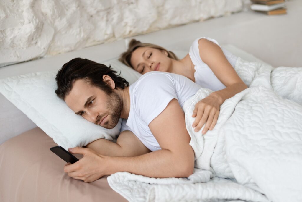 Angry man checking his sleeping girlfriend's smartphone, lying in bed at home, husband feeling