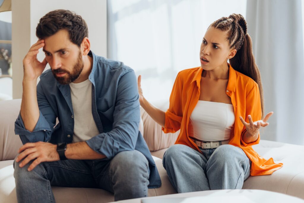 Portrait of upset man being yelled at by angry, abusive woman, sitting on sofa in room at home