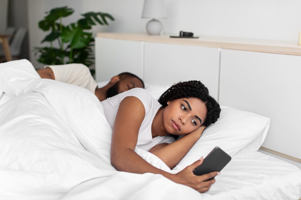 Sad young african american lady looks at smartphone lie on comfortable bed hide cheating