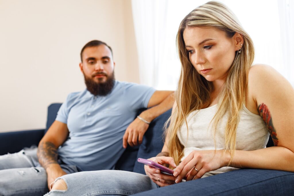 Suspicious man looking at his girlfriend's smartphone screen while they sit on sofa